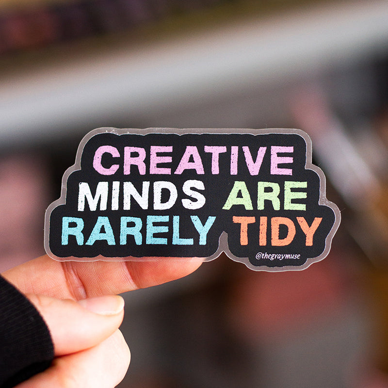 Hand holds a clear matte sticker with multicolor text reading Creative Minds Are Rarely Tidy against a dark backdrop.