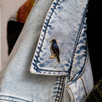 American Crow enamel pin with a gold edge attached to light wash denim jacket