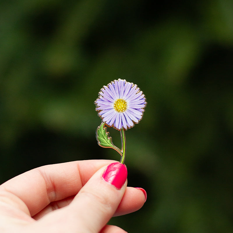 Purple aster enamel pin held by fingers against a blurred green background