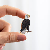 Hand holds Bald Eagle enamel pin with white head and dark body against a light background.