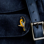 Baltimore Oriole enamel pin with orange and black feathers pinned to dark denim beside a brass belt buckle