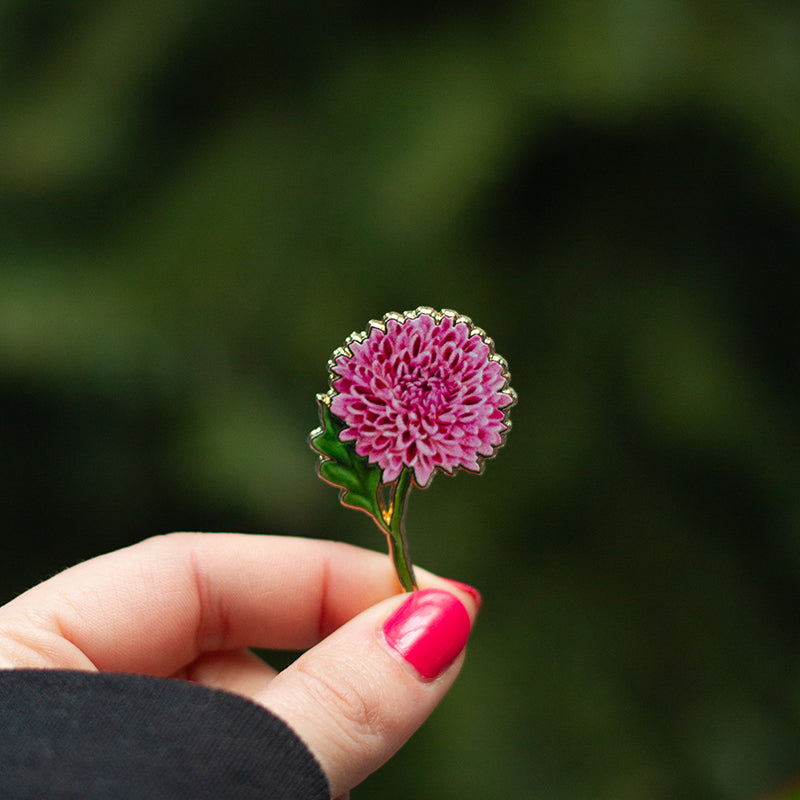 Close-up of a pink chrysanthemum enamel pin held by fingers against a blurred green background.