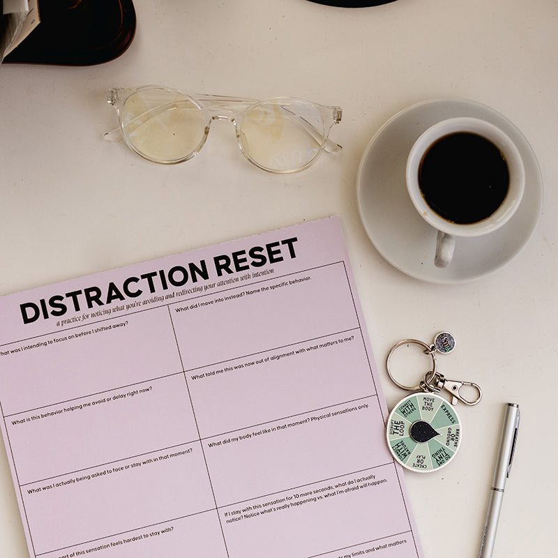 Overhead view of a pale pink distraction reset notepad on a white desk with glasses, a coffee cup, and a spinner keychain.