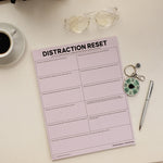 Top view of a pink DISTRACTION RESET notepad with coffee, glasses, and a spinner keychain.
