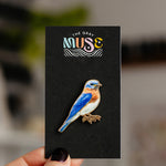 Colorful bluebird enamel pin mounted on a black display card with The Gray Muse logo.
