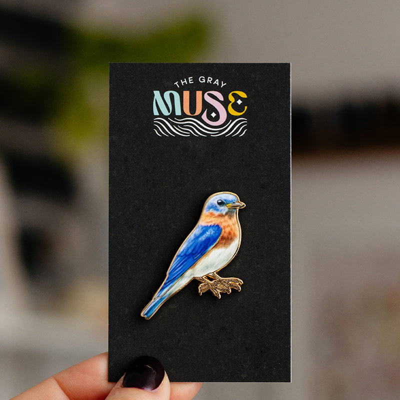 Colorful bluebird enamel pin mounted on a black display card with The Gray Muse logo.