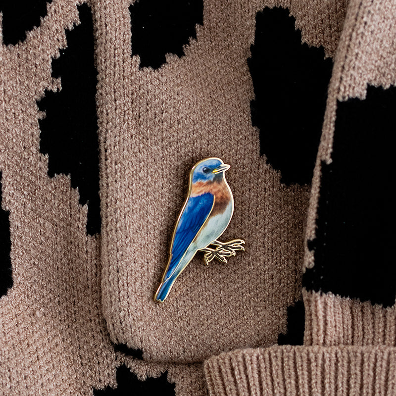 Eastern Bluebird enamel pin with bright blue wings and russet chest resting on a brown knit sweater.