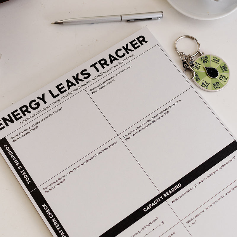 Energetic Capacity Bundle on a white desk with the Energy Leaks Tracker notepad, a silver pen, and a green spinner keychain.