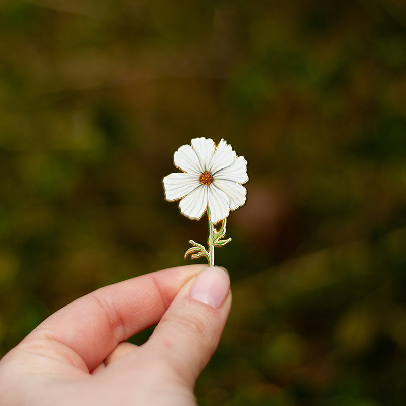 White Cosmos enamel pin held between fingertips against a blurred outdoor background.