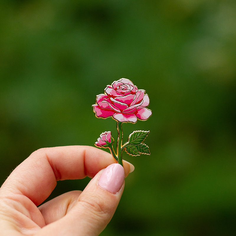 Pink rose enamel pin held by fingers against a soft green background