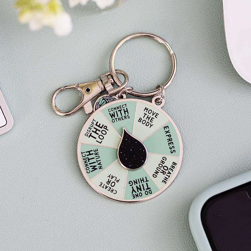 Close up of Get Unstuck spinner enamel keychain with mint green segments and a black teardrop center on a soft blue surface
