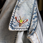 Enamel pin of a green hummingbird clearwing moth with red wing accents, pinned to a light denim jacket cuff.