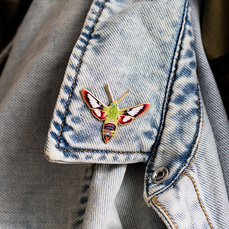 Enamel pin of a green hummingbird clearwing moth with red wing accents, pinned to a light denim jacket cuff.