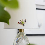 Hummingbird Clearwing Moth enamel pin placed on a white lampshade beside a faceted crystal lamp base.