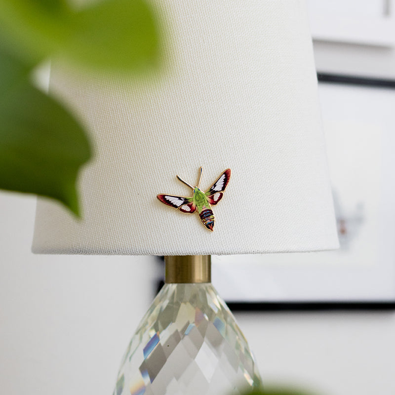 Hummingbird Clearwing Moth enamel pin placed on a white lampshade beside a faceted crystal lamp base.