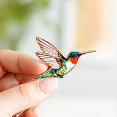 Colorful hummingbird enamel pin held between fingers against a soft blurred background