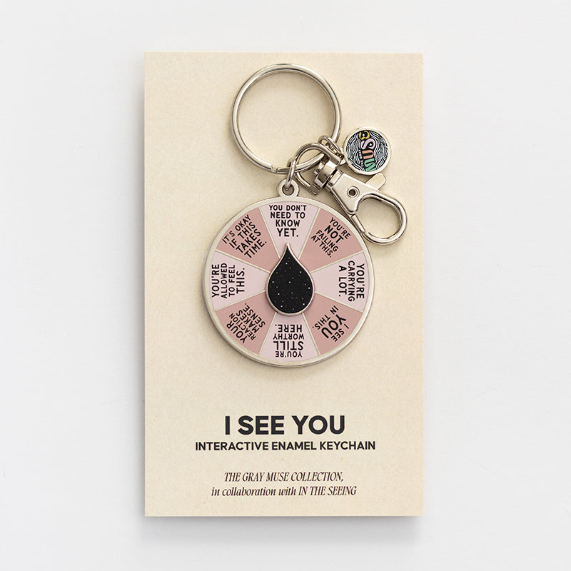 I See You Spinner Interactive Enamel Keychain on a beige display card with a pink affirmation wheel and a black teardrop centerpiece