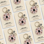 Assorted I See You spinner enamel keychains on beige display cards.