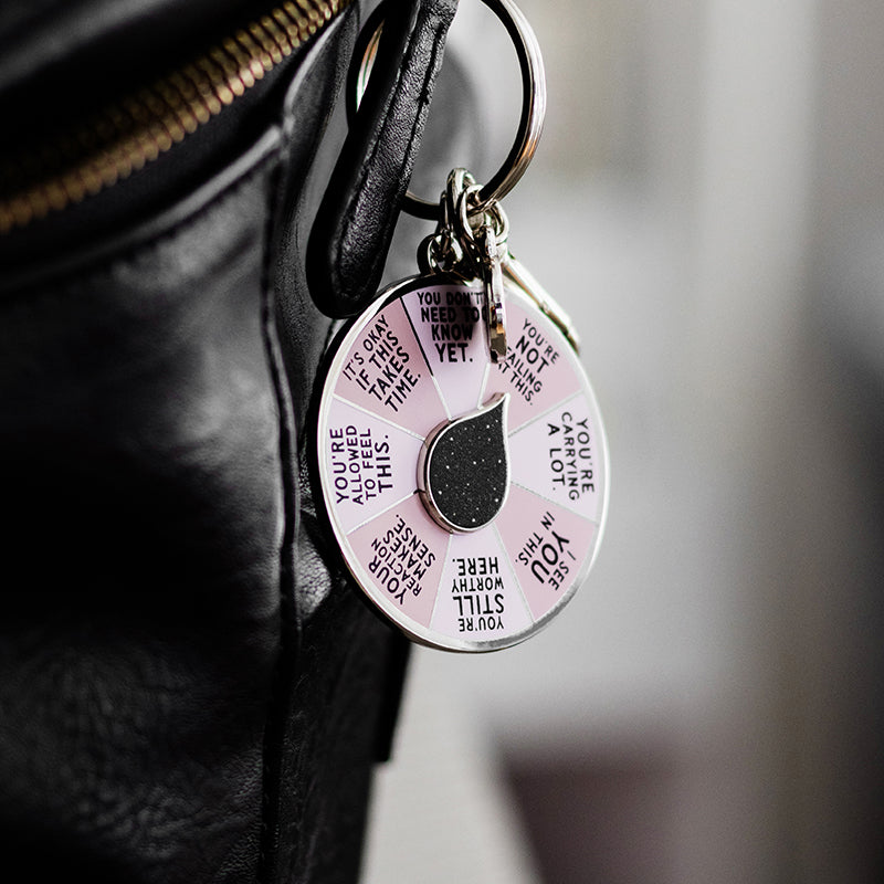 Close up of I See You Spinner Interactive Enamel Keychain attached to a black bag, showing a pink affirmation wheel and a colorful circular charm