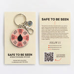 Front and back display of Safe to Be Seen spinner enamel keychain on beige cards; front shows the keychain with pink wheel, back has product description.
