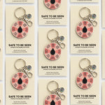 Grid of Safe to Be Seen spinner enamel keychains on beige display cards, each with a pink segmented wheel and a black teardrop center.