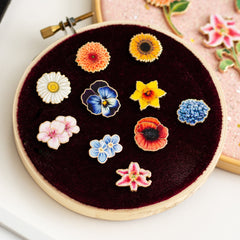 Ten miniature floral enamel pins arranged on burgundy velvet inside a round hoop display