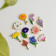Ten miniature floral enamel pins from Mini Floral Enamel Pin Collection Set 3 arranged on a white background including Indian Paintbrush, Jonquil, California Poppy, Cosmos, Morning Glory, Purple Orchid, Violet, Magnolia, Pink Rose, and Wisteria.