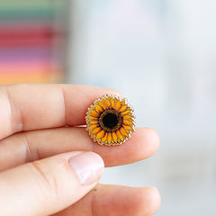 Mini sunflower enamel pin held between fingertips with bright yellow petals and dark center
