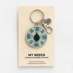 My Needs interactive enamel keychain displayed on a beige card with the spinning wheel and attached charm.