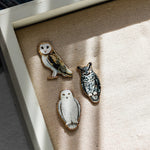 Three owl enamel pins arranged on a textured beige fabric surface with soft shadows.