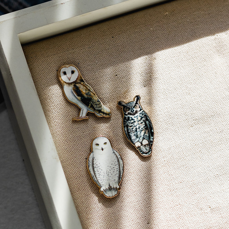 Three owl enamel pins arranged on a textured beige fabric surface with soft shadows.
