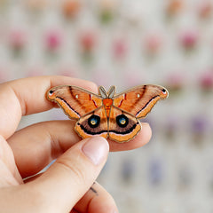 2.1 inch Polyphemus Moth enamel pin held between fingers against a blurred background