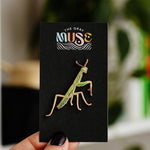 Green and gold praying mantis enamel pin mounted on a black display card with The Gray Muse logo.