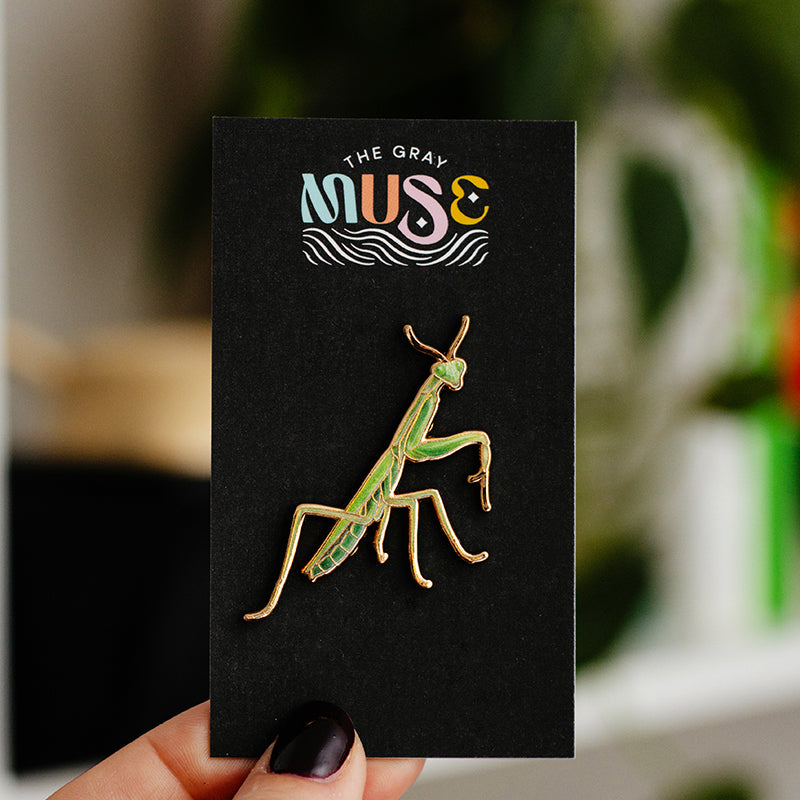 Green and gold praying mantis enamel pin mounted on a black display card with The Gray Muse logo.