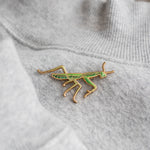 Praying Mantis enamel pin on gray fabric with green and gold enamel detailing.