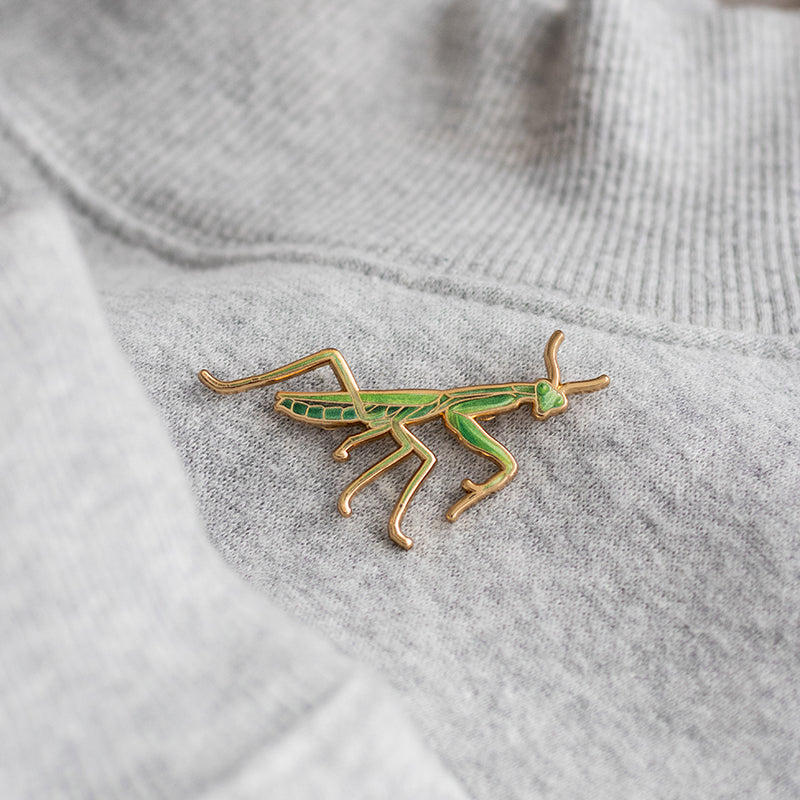 Praying Mantis enamel pin on gray fabric with green and gold enamel detailing.