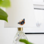 Red Admiral enamel pin with dark wings and orange bands on a white lampshade.