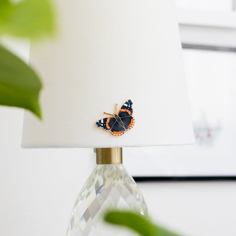 Red Admiral enamel pin with dark wings and orange bands on a white lampshade.