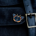 Red Admiral enamel pin on dark denim fabric beside a brass belt buckle.