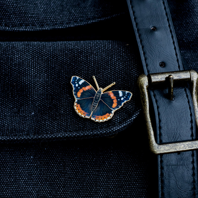 Red Admiral enamel pin on dark denim fabric beside a brass belt buckle.