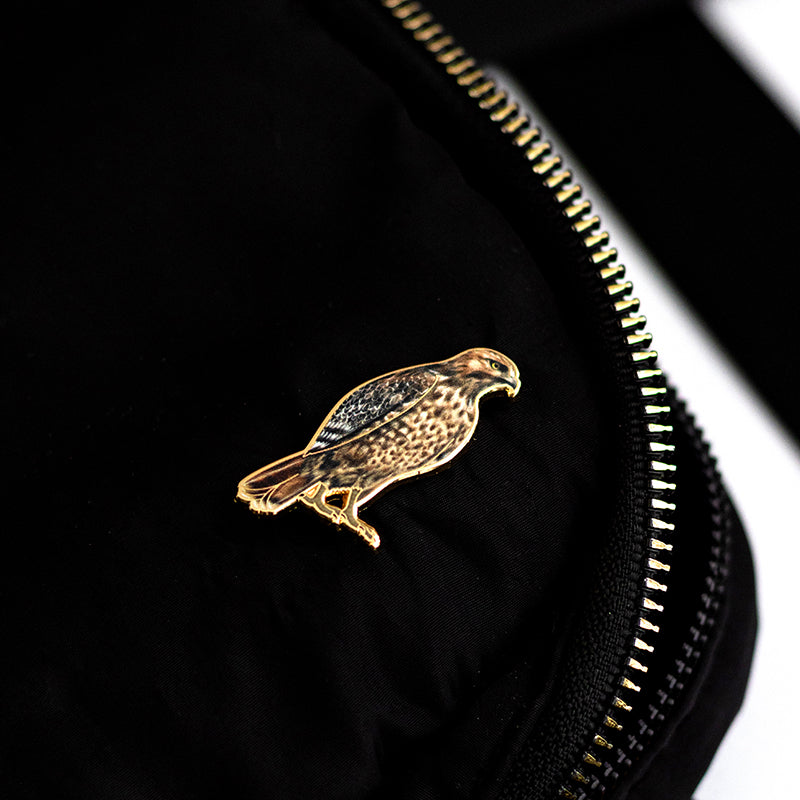 Red-tailed hawk enamel pin perched on black fabric beside a gold zipper