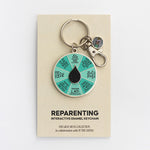 Circular enamel keychain with teal segmented spinner and a black teardrop center on a beige display card.