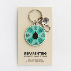 Circular enamel keychain with teal segmented spinner and a black teardrop center on a beige display card.