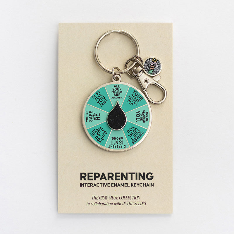 Circular enamel keychain with teal segmented spinner and a black teardrop center on a beige display card.