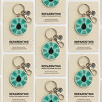 Reparenting interactive enamel keychain with teal segmented spinner and metal ring on a beige card.