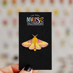 Rosy Maple Moth enamel pin with pink and yellow wings mounted on a black display card.