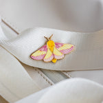 Rosy Maple Moth enamel pin featuring pink and yellow wings resting on a white ribbon against a light background.