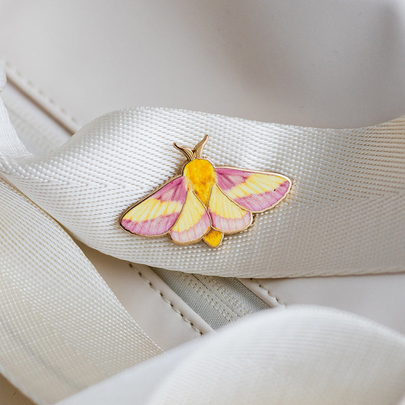 Rosy Maple Moth enamel pin featuring pink and yellow wings resting on a white ribbon against a light background.