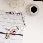 Safe to be Seen Bundle on a white desk with Identity Shift Notepad, spinner, expansion edge slider keychain, coffee, and glasses.
