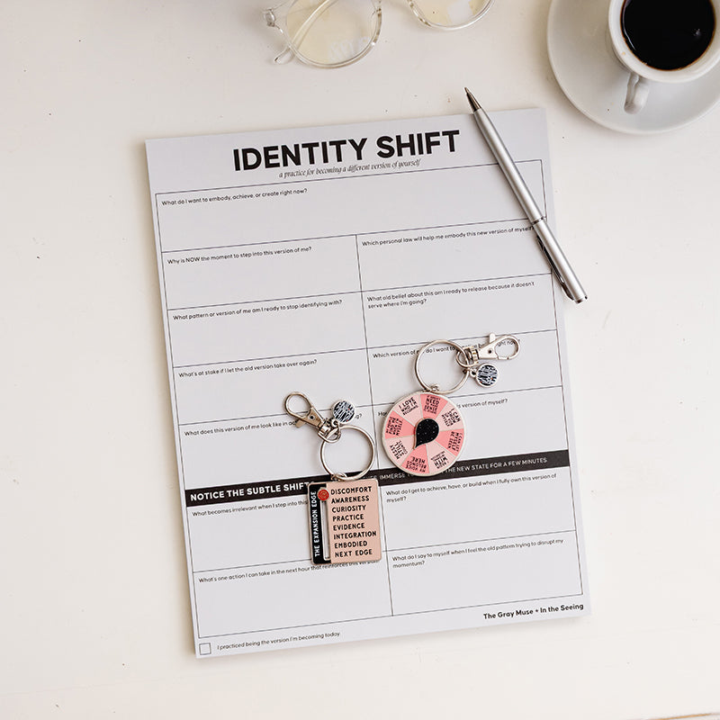 Identity Shift notepad on a white desk with a silver pen, clear glasses, coffee, and two small keychain charms.